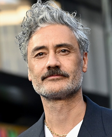 Taika Waititi headshot wearing white shirt with a black suit jacket and gold chain necklace.