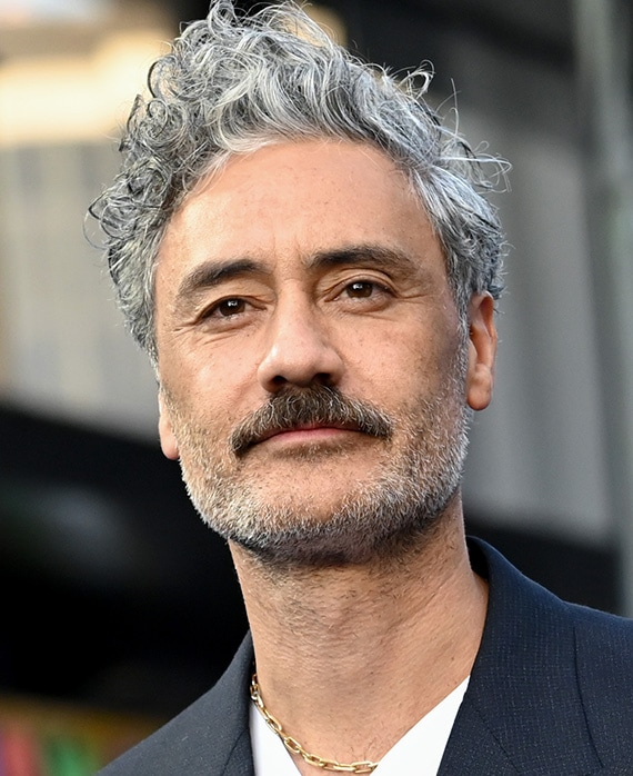 Taika Waititi headshot wearing white shirt with a black suit jacket and gold chain necklace.