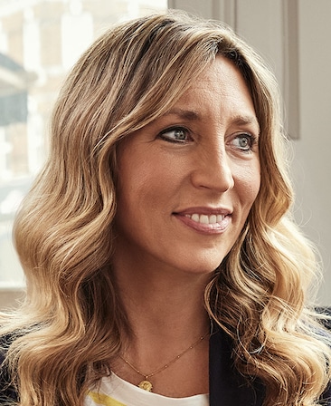 Daisy Haggard headshot wearing a gold necklace with a white shirt and black jacket.
