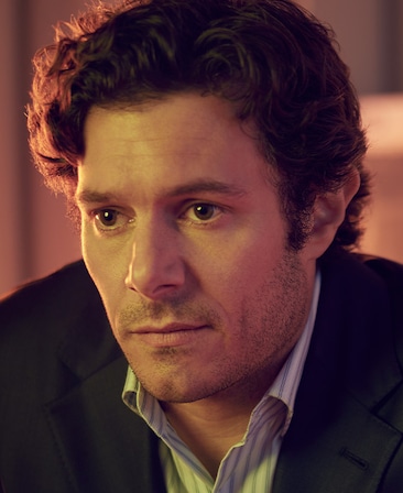 Adam Brody headshot in black coat with blue and white striped button down, with half face illuminated by peach colored light