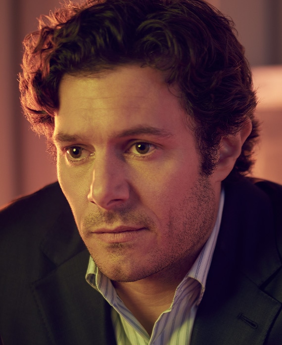 Adam Brody headshot in black coat with blue and white striped button down, with half face illuminated by peach colored light