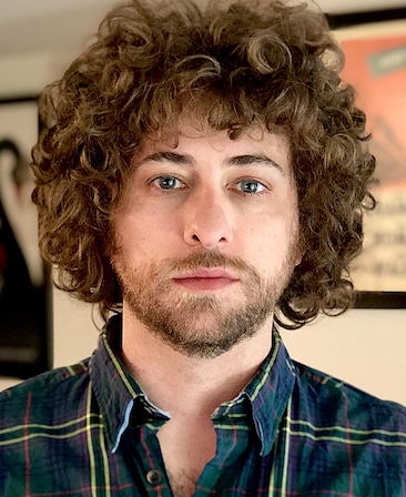 Kieran Valla headshot wearing a green and yellow plaid shirt with pictures hanging in the background