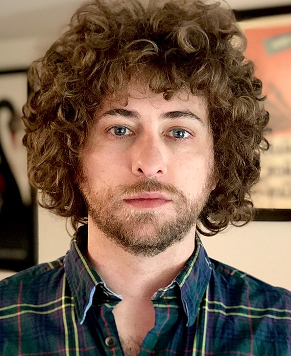 Kieran Valla headshot wearing a green and yellow plaid shirt with pictures hanging in the background