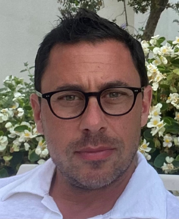 Headshot of executive producer Nicholas Frenkel, wearing black glasses and a white button up in front of flowered greenery.