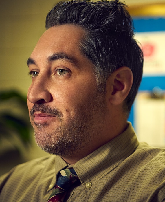 Carmen Christopher as Rick for FX's English Teacher wearing a green plaid button up and multi-colored tie facing to his side.