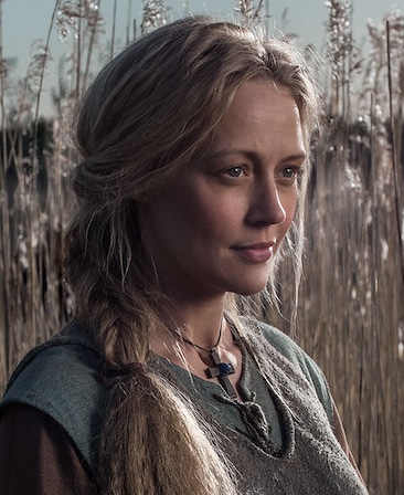 Elen Rhys headshot standing in wheat field with peasant blue and grey layered clothes with wooden cross necklace