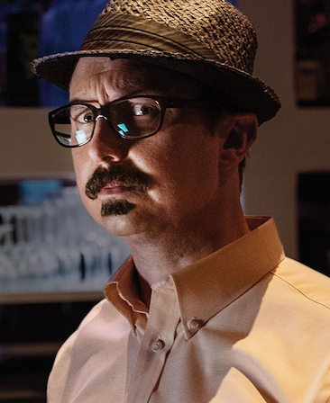 John Hodgman headshot wearing black glasses, a fedora hat and orange button up shirt
