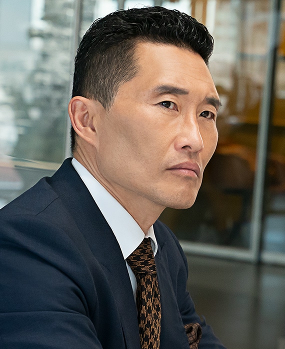 Daniel Dae Kim headshot wearing a navy suit jacket and orange patterned tie, standing in front of a window