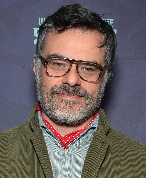 Jemaine Clement headshot wearing glasses with a green jacket.