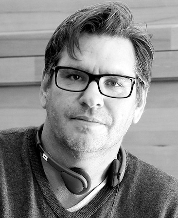 Black and white headshot of Dario Scardapane in v neck sweater with headphones around neck and light wood wall background