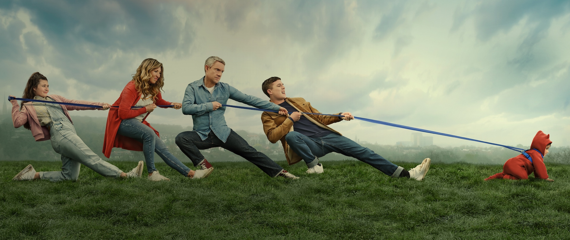 Cast of Breeders family amid a grassy field pulling on a rope tethered to a crawling baby dressed in a red onesie