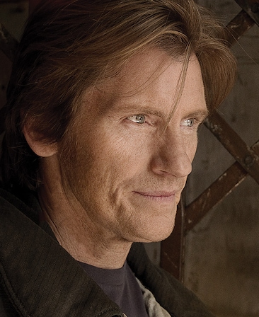 Denis Leary headshot wearing a black shirt and standing by a metal fence