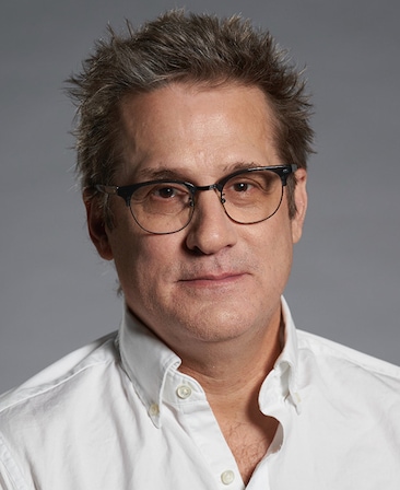 Headshot of executive producer Paul Simms wearing a white button up, with a grey background.