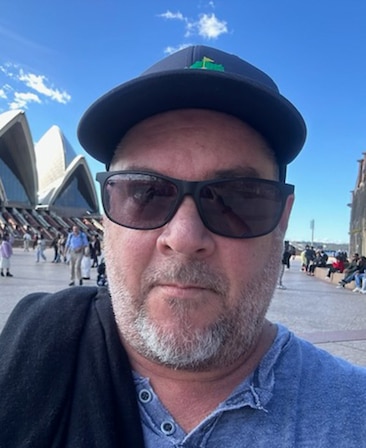 Josh Drisko headshot wearing a black hat and sunglasses standing outside of the Sydney Opera House