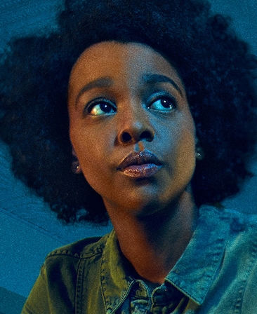 Mallori Johnson grainy filtered headshot in collared blue denim top, looking up toward soft light shining down on face