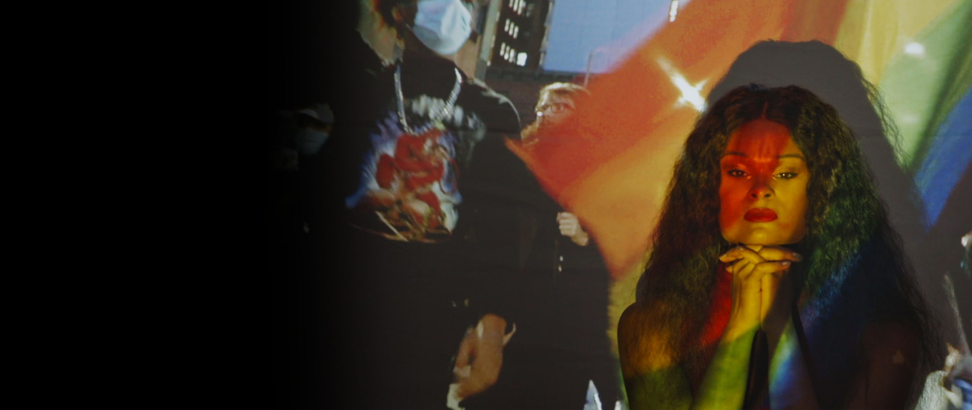 Woman in front of a projector displaying a PRIDE parade with masked participants, and the rainbow flag colors on her face