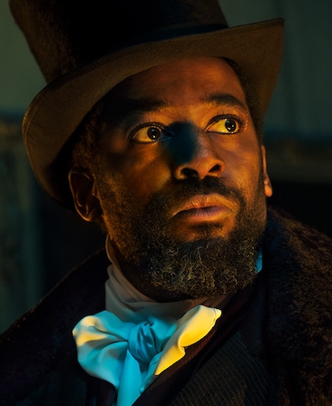 Ashley Thomas headshot in black coat with white silk neck tie scarf with black top hat, looking up toward warm light