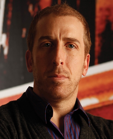 Todd Kessler headshot in blue and red striped button down with red and white image background and half his face shadowed