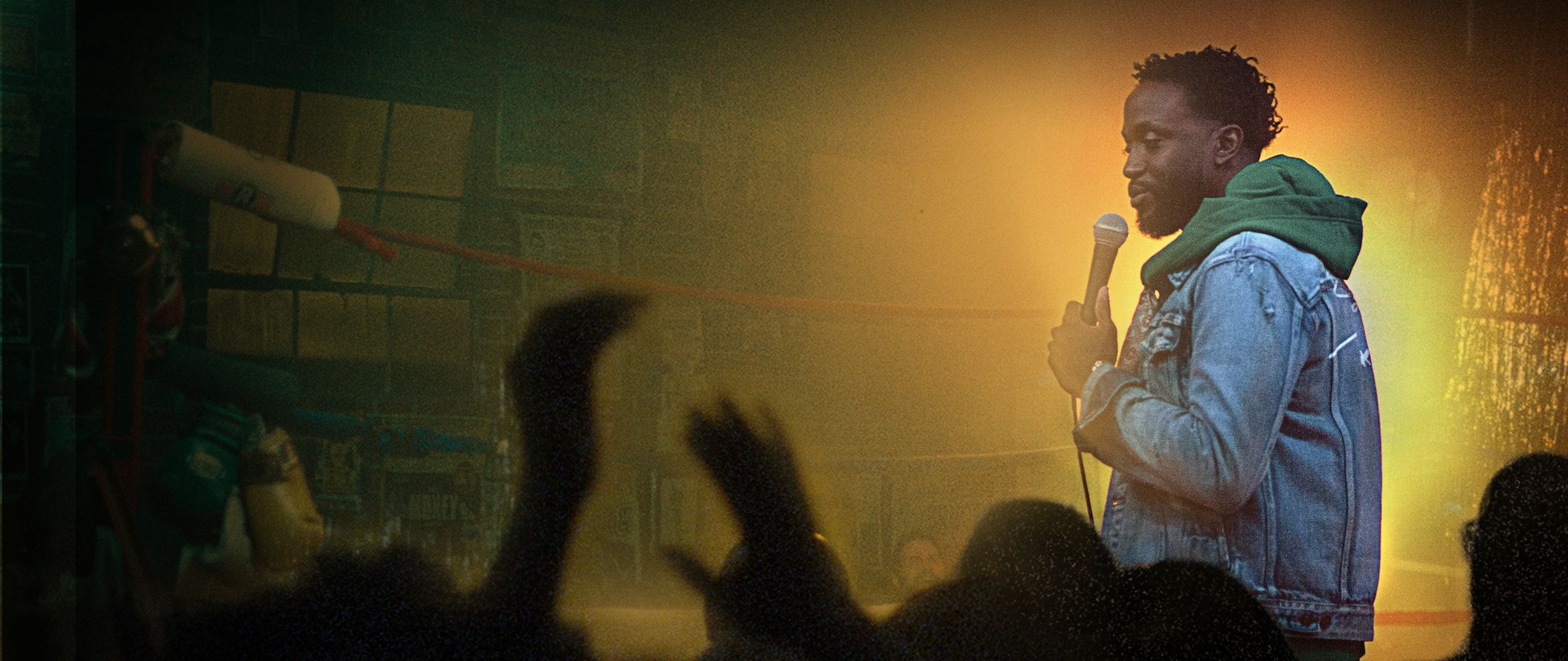 Comedian Byron Bowers on a stage holding a mic wearing a denim jacket and green hoodie with a spotlight shining behind him