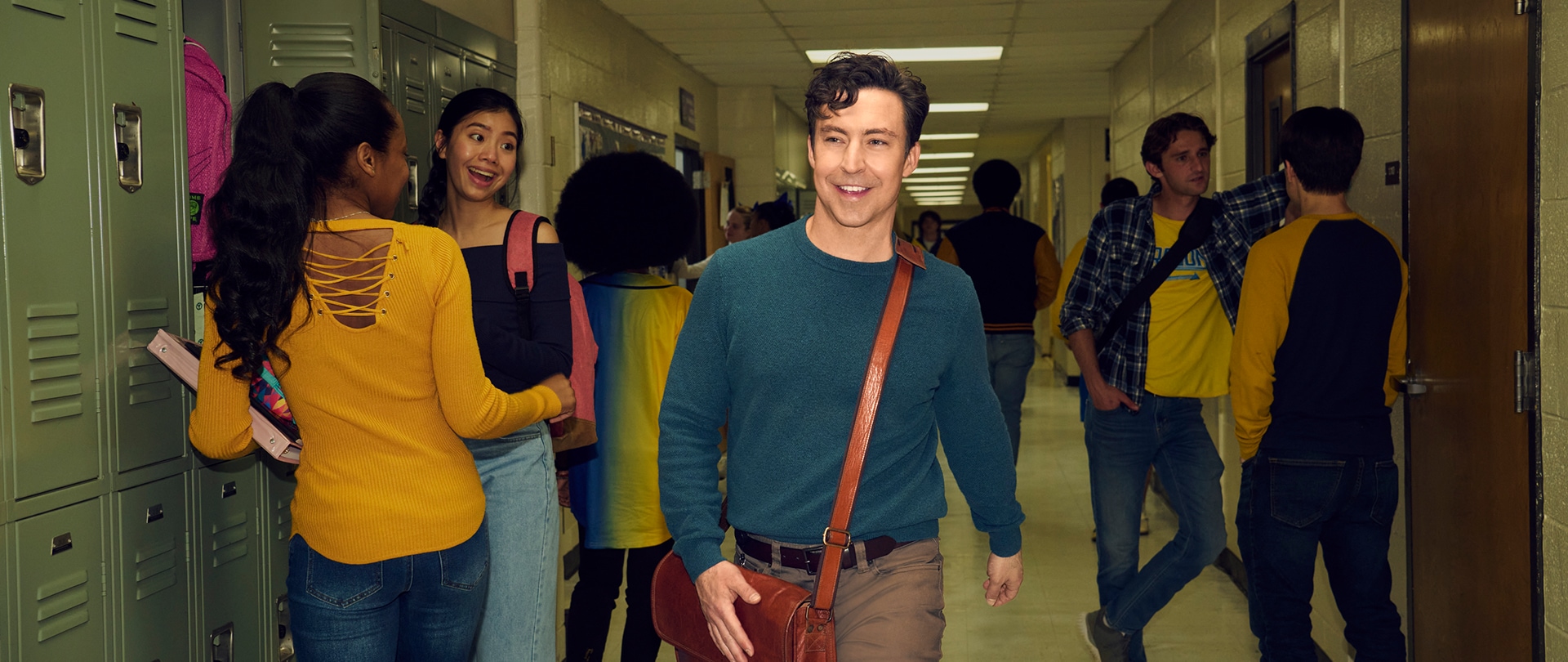 Evan Marquez wearing a teal sweater from FX's 'English Teacher' walking down a busy school hallway full of students.