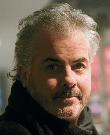 Peter Ofallon headshot wearing a black scarf and standing in front of a window