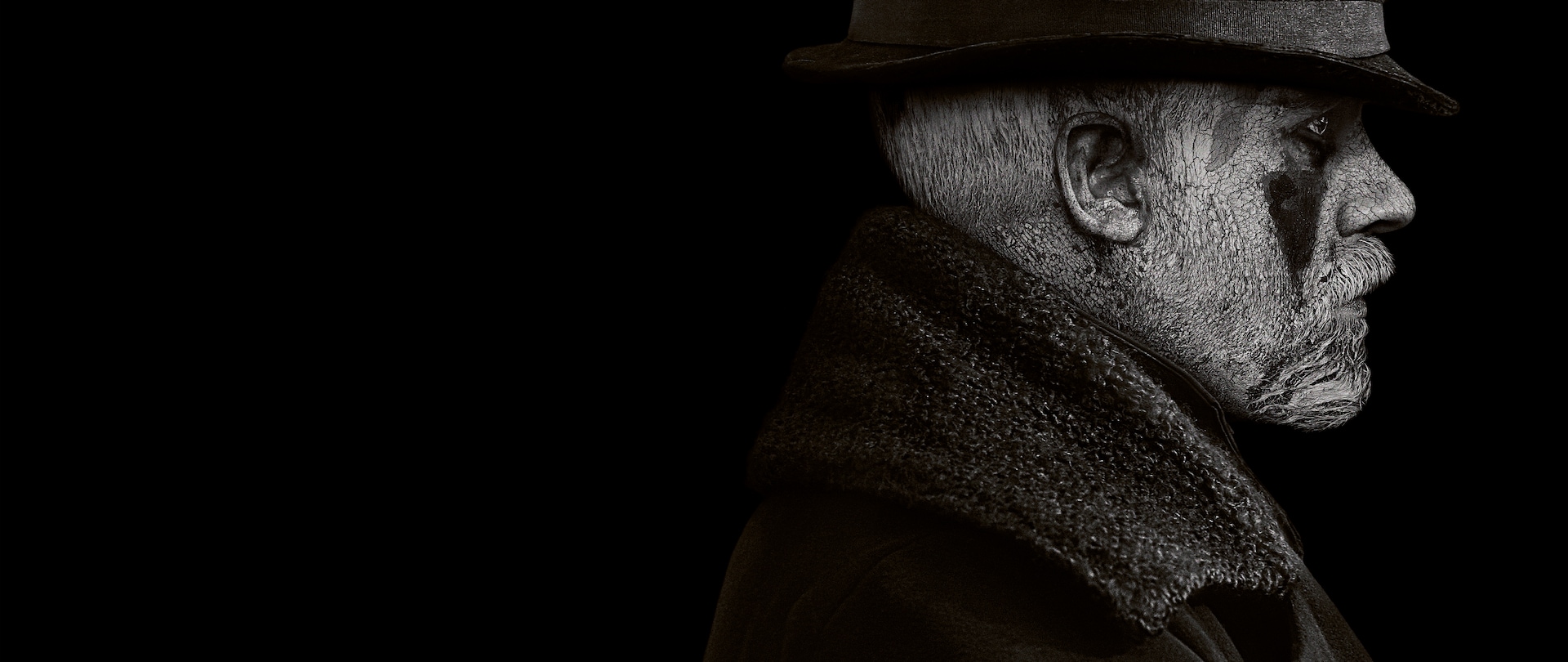 Profile of Tom Hardy with face painted white, reverted black triangles under the eyes, wearing black hat and black wool coat