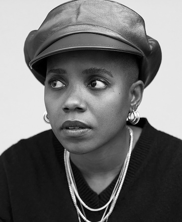 Janzica Bravo black and white headshot in leather cap, black v neck sweater, and wearing hoop earrings and necklaces
