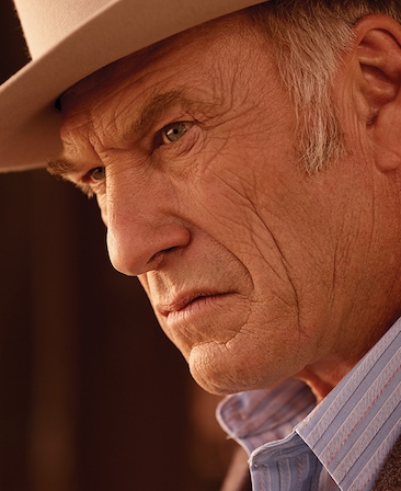 Ted Levine headshot in light blue striped button down and wearing white top hat with sun shining on face