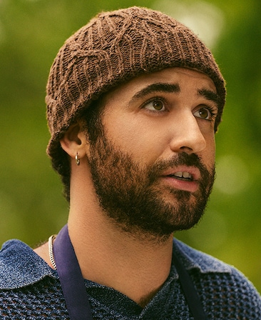 Jordan Firstman as Malcolm in FX's English Teacher, wearing a brown beanie, textured collared shirt and blue apron outdoors.