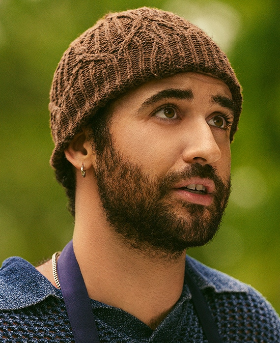 Jordan Firstman as Malcolm in FX's English Teacher, wearing a brown beanie, textured collared shirt and blue apron outdoors.