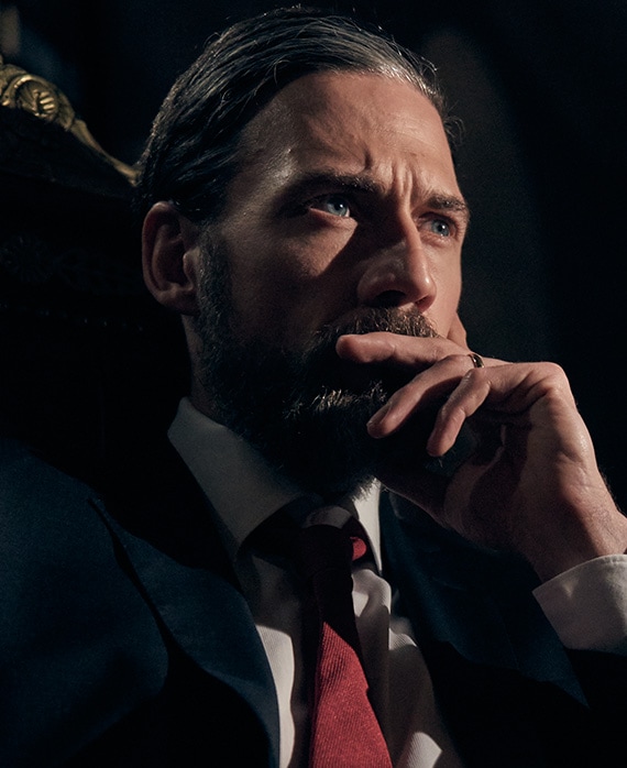 Adam Rayner headshot wearing a suit and placing his hand on his month