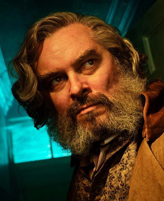Trystan Gravelle headshot in brown coat with golden embellished vest, looking up toward bright light in dark room with window