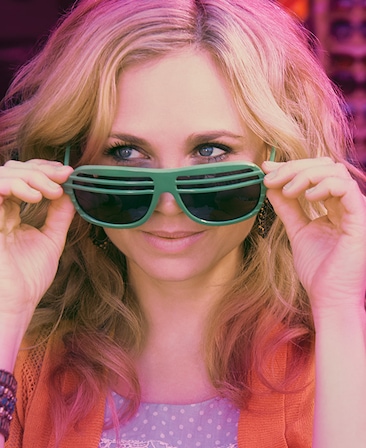 Fiona Gubelmann headshot wearing green sunglasses, a polka dot top and orange scarf
