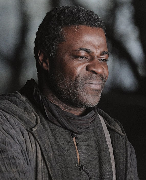 Danny Sapani headshot in ancient peasant black robe clothing with shadows of branches in background