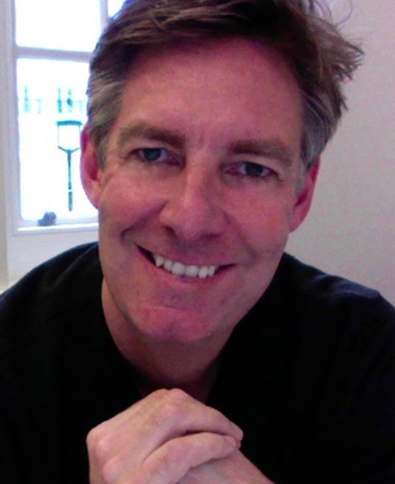 Dean Baker headshot in plain black t shirt with bright window and white wall in background