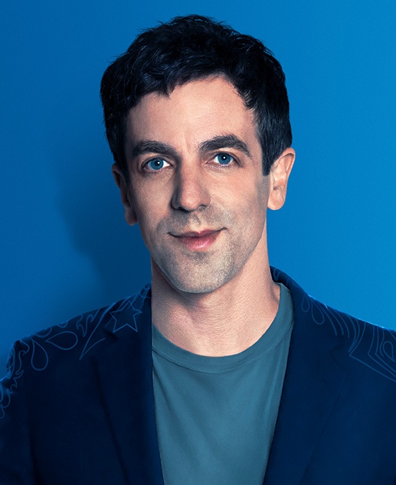 B.J. Novak headshot wearing a teal shirt, navy jacket and standing in front of a blue wall