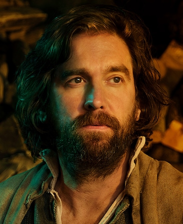 Owen McDonnel headshot in brown jacket with half face illuminated by green light and other brightly illuminated by warm light