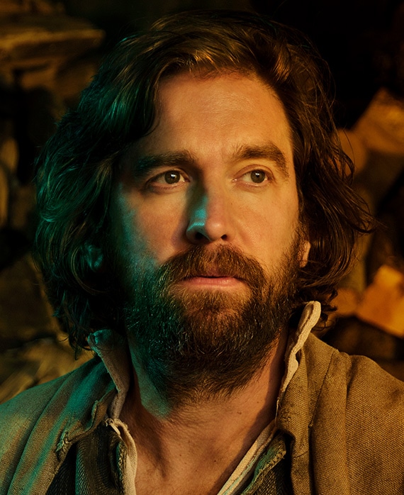 Owen McDonnel headshot in brown jacket with half face illuminated by green light and other brightly illuminated by warm light