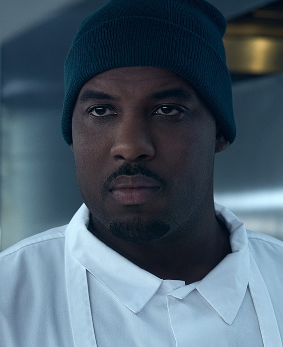 Lionel Boyce headshot wearing blue beanie and white collared shirt and apron.