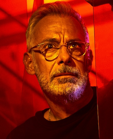 Headshot of Joe Mantello wearing clear glasses and black shirt by red lighting