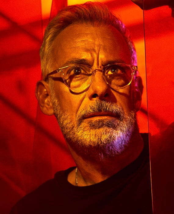 Headshot of Joe Mantello wearing clear glasses and black shirt by red lighting