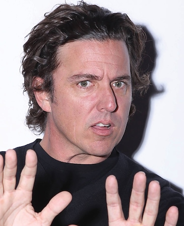 Chris Case headshot in plain black t shirt with bright light casting shadow on white background, with both hands up