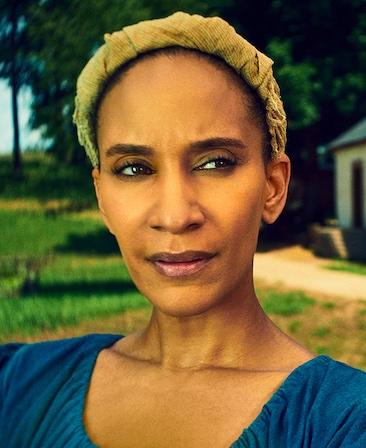 Sophina Brown headshot in blue peasant top with yellow headband, with grass and trees in background with side of white shed