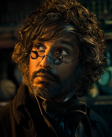 Rudi Dharmalingam headshot in dark brown coat with bowtie neck scarf, wearing metal spectacles and looking at bright light