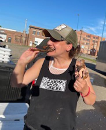Executive producer Kathryn Dean in a black tank top and green hat, outside eating a handful of chicken drumsticks.