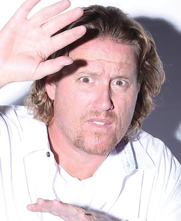 Paul Bernard headshot in white shirt with bright light shining on face and casting shadow on white wall behind, with hands up