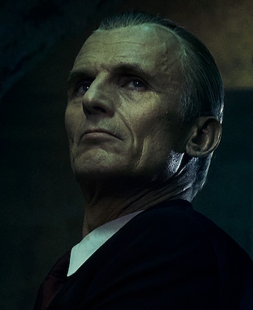 Richard Sammel Headshot wearing a black jacket with white collar and staring above 