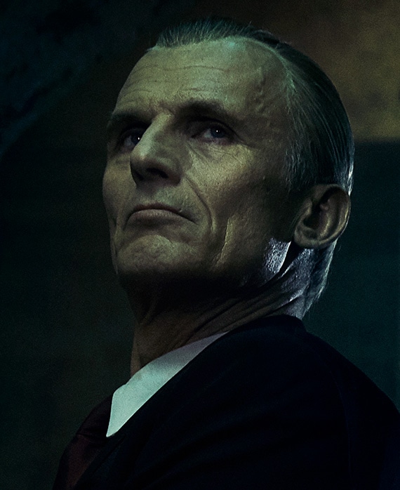 Richard Sammel Headshot wearing a black jacket with white collar and staring above 