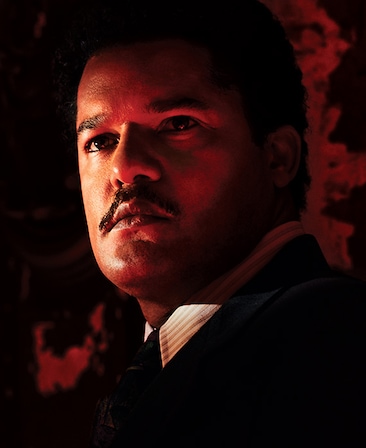 Brandon J. Dirden headshot in black suit over white striped collar, looking up with half his face illuminated in red lighting