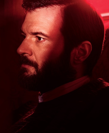Costa Ronin headshot in dark room, in black suit and white collared button down, with half his face illuminated in red light
